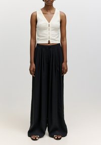 Beige Cropped-Top mit V-Ausschnitt und Knopfdetail; kombiniert mit lockeren, schwarzen, plissierten Hosen. Sandalen mit dekorativen Akzenten.