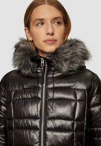 Schwarze Daunenjacke mit glänzender Oberfläche und gestepptem Design. Verfügt über einen abnehmbaren grauen Pelzkragen und einen vorderen Reißverschluss mit Kordelzügen.