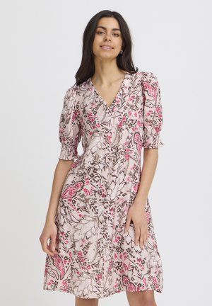 Žena v kvetinových šatách po kolená s nafúknutými rukávmi a výstrihom do V, stojaca pred jednoliatym svetlým pozadím.