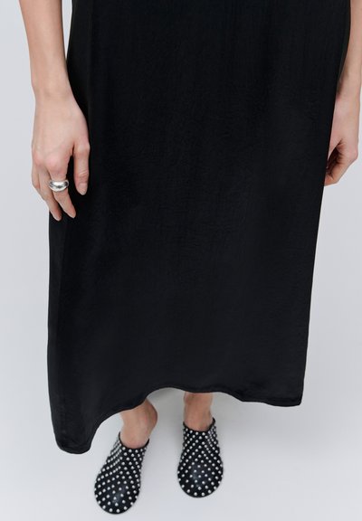 Personne portant une longue robe noire, des chaussures noires à enfiler avec des clous blancs, et une bague argentée au majeur de la main gauche.