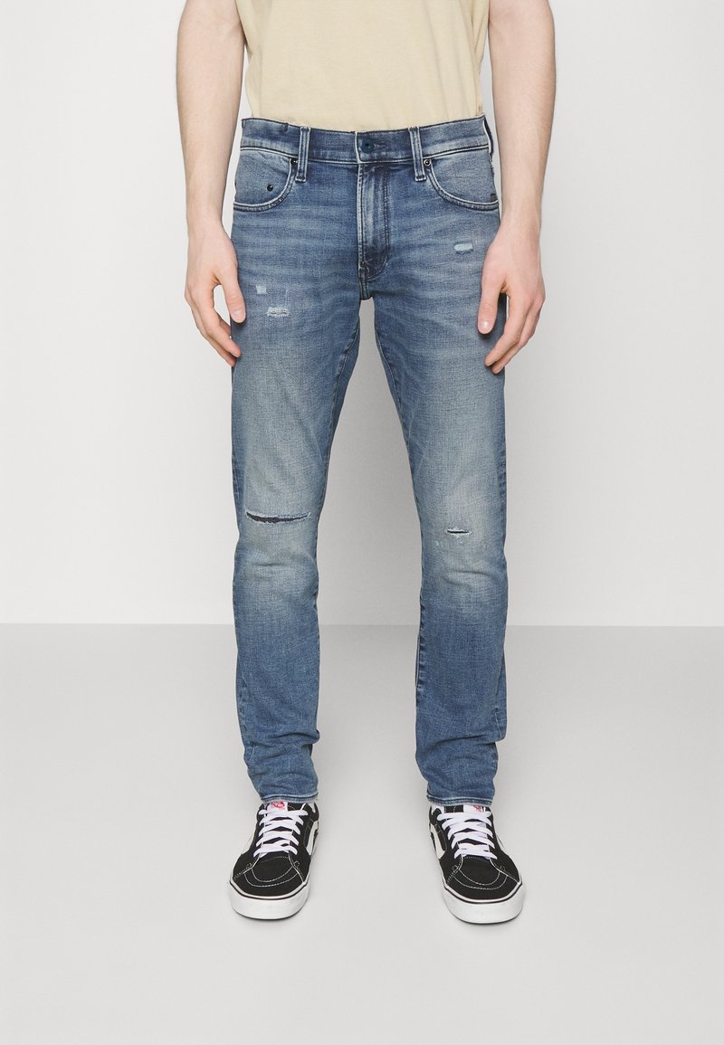 Hombre vistiendo jeans ajustados desgastados de color azul y zapatillas de laceado en blanco y negro, de pie contra un fondo claro y sencillo.