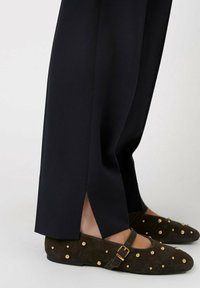 Black tailored trousers with side slits, paired with dark brown suede shoes featuring gold studs and a strap across the foot.