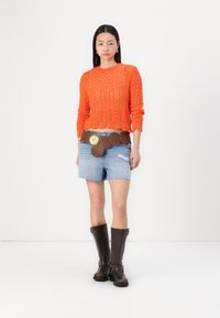 Maglione arancione a maglia con scollatura rotonda, pantaloni corti in denim blu, cintura marrone con dettagli circolari e stivali alti fino al ginocchio in pelle marrone.