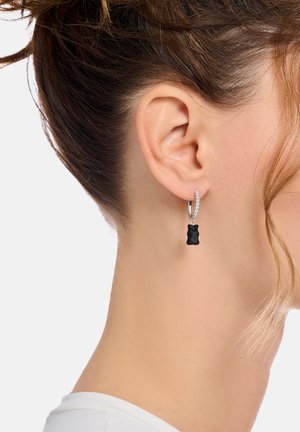 Schwarzer Gummibärchenanhänger, der von einem silbernen Creolenohrring herabhängt, verziert mit kleinen klaren Edelsteinen, getragen am Ohr.