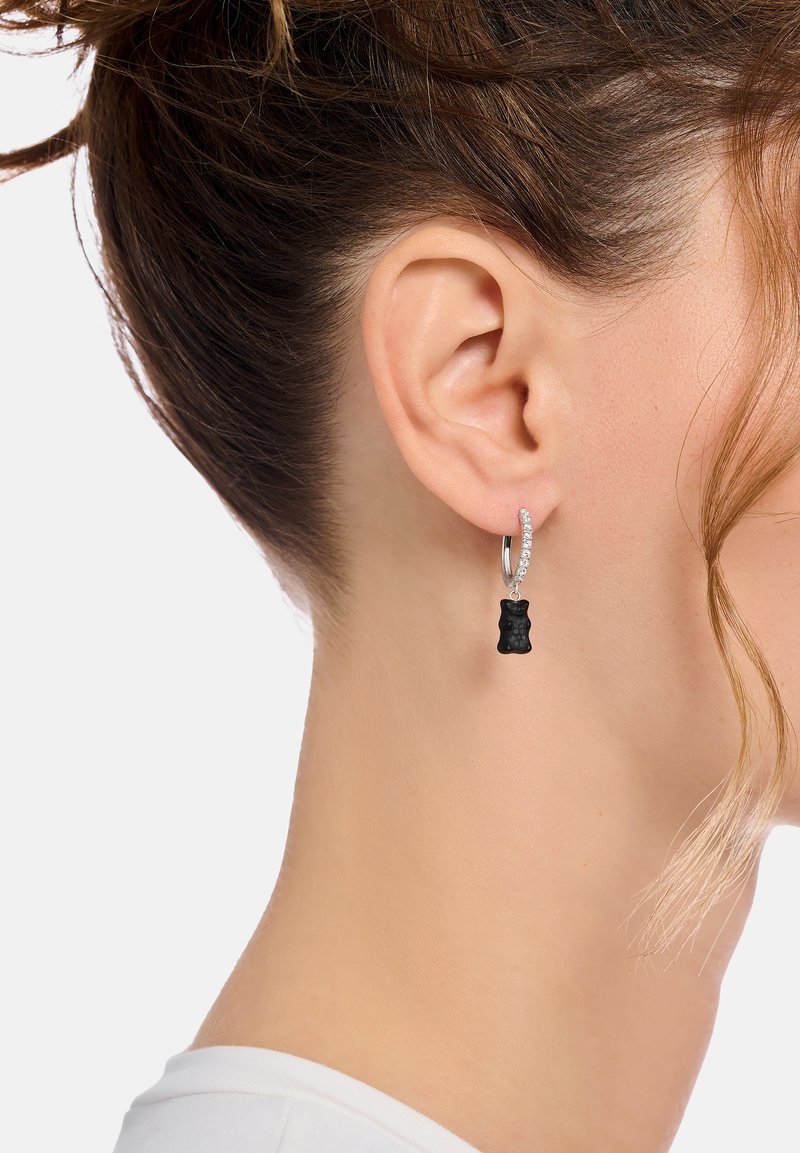 Schwarzer Gummibärchenanhänger, der von einem silbernen Creolenohrring herabhängt, verziert mit kleinen klaren Edelsteinen, getragen am Ohr.