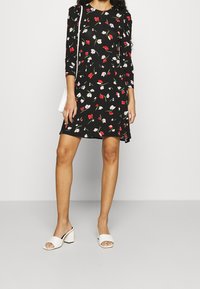 Kobieta nosząca czarną sukienkę w kwiaty z rękawami do trzech czwarte, białe sandały na obcasie i małą białą torebkę na ramię.