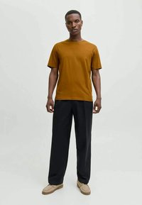 Uomo in piedi che indossa una maglietta marrone senape, pantaloni neri larghi e scarpe beige su uno sfondo chiaro e semplice.
