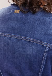 Mörkblå denimjacka med en urblekt textur, klassisk design, synliga sömmar och en liten gul bröjtagg på baksidan av kragen.