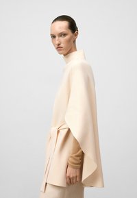 Zacht perzikkleurig poncho-stijl top met een hoge kraag, gedrapeerd ontwerp en een strikaccent in de taille, gemaakt van een gestructureerde, lichte stof.