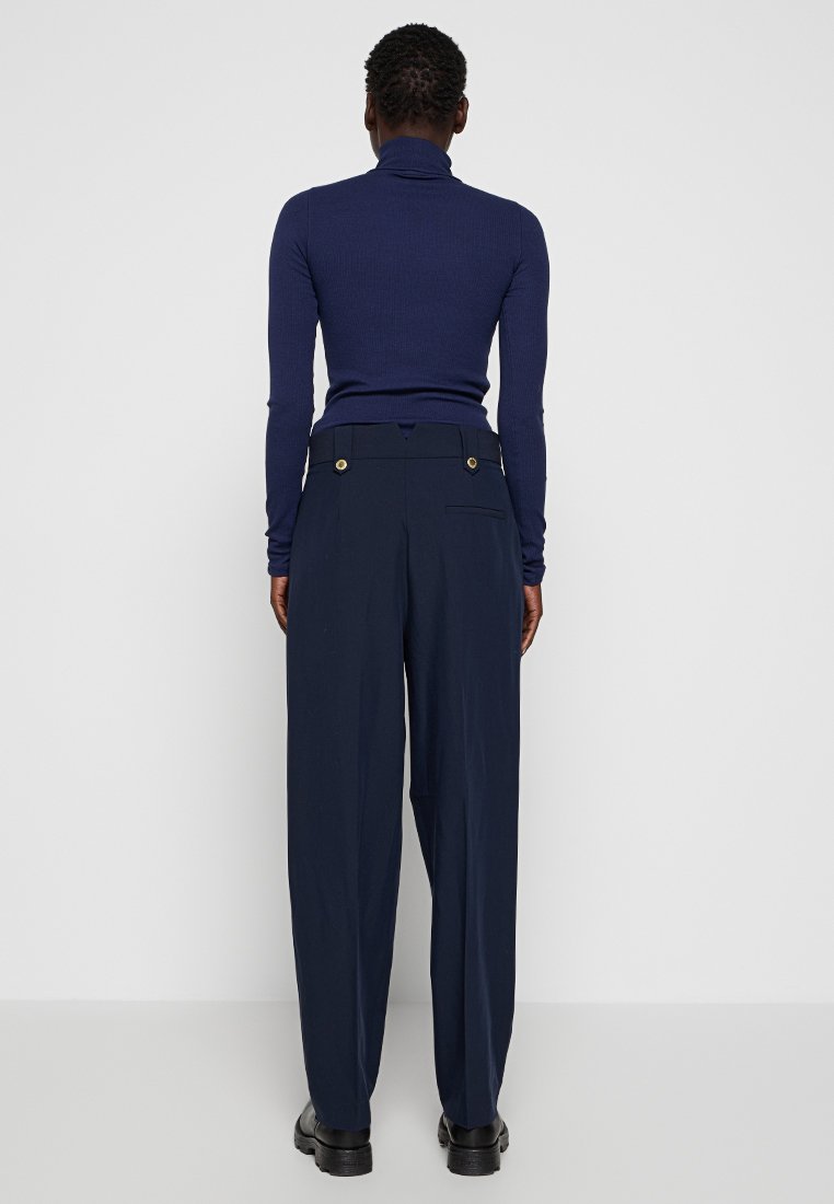 Personne portant un col roulé bleu marine et un pantalon bleu marine taille haute avec des détails de boutons, se tenant face à un mur blanc.