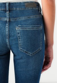 Dark blue denim jeans feature a mid-rise waistband, plain back pockets, and a leather brand patch at the top center of the waistband.