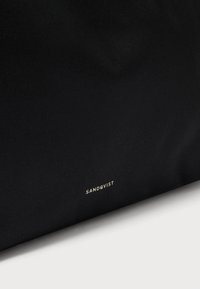 Juodas tekstilinis krepšys, turintis struktūruotą dizainą, subtilią faktūrą ir metalinį auksinį „SANDQVIST“ logotipą prie apačios.