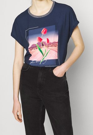 Osoba ubrana w granatowy t-shirt z różowymi tulipanami i grafiką pustynnego krajobrazu, w połączeniu z czarnymi spodniami z wysokim stanem, ręka w kieszeni.