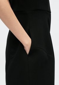 Μαύρο φόρεμα με υφή υφάσματος, τσέπη στο πλάι και εφαρμοστή σιλουέτα, με καθαρές γραμμές και διακριτή ματ φινίρισμα.