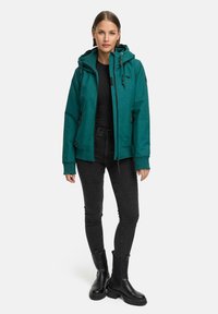 Teal vanntett jakke med hette, glidelås foran og justerbare mansjetter. Brukt over en svart topp og kombinert med svarte jeans og støvler.