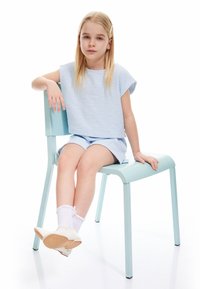 Hellblaues, strukturiertes, kurzes Oberteil und Shorts, sitzend auf einem mintgrünen Stuhl. Weiße Schuhe und Socken mit Rüschen, glattes, blondes Haar.