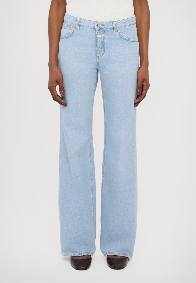 Personne portant un jean bleu clair taille haute et jambes larges avec des chaussures noires, debout les bras détendus le long du corps sur un fond uni.