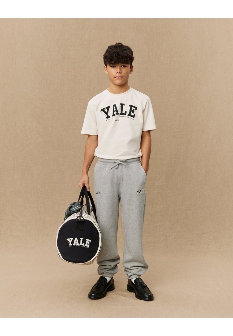 Weißes T-Shirt mit dem Aufdruck "YALE" in Schwarz, graue Jogginghose mit Logo, schwarze Schuhe, hält eine runde schwarze Sporttasche mit "YALE"-Logo.