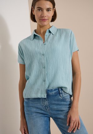 Frau mit kurzen braunen Haaren trägt ein hellblaues kurzärmliges Hemd und blaue Jeans und steht vor einem schlichten Hintergrund.