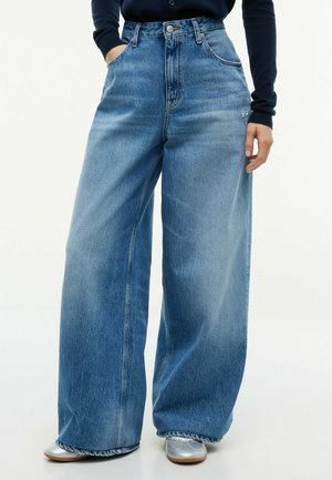 Personne portant un jean bleu taille haute à jambes larges, les mains dans les poches et des chaussures argentées, debout devant un fond clair uni.