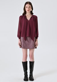 Blusa di seta bordeaux con un design fluido, caratterizzata da un collo a V e maniche lunghe, abbinata a una mini gonna rosa testurizzata e stivali neri fino al ginocchio.