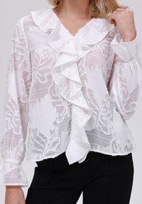 Hvit gjennomsiktig blouse med blomstermønster, volangkrage og front, lange ermer med elastiske mansjetter, og en avslappet passform.