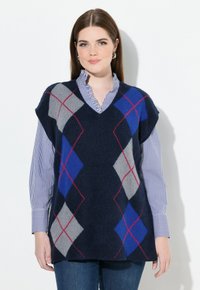 Mladá žena nosící stříbrné kruhové náušnice, námořnickou argyle vestu přes modro-bíle pruhovanou košili a modré džíny, jemně se usmívající.