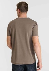 Uomo che indossa una maglietta a maniche corte marrone semplice e pantaloni scuri, in piedi con la schiena rivolta verso uno sfondo chiaro e neutro.