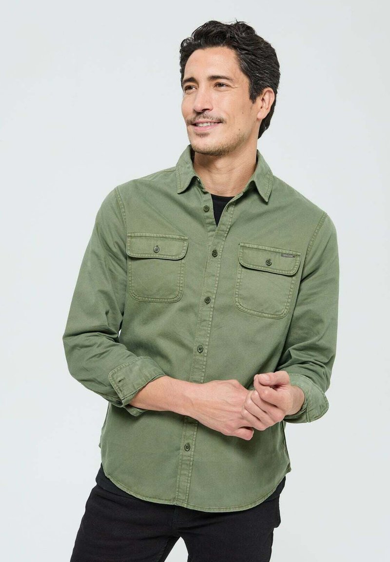 Chemise en coton vert olive à manches longues, avec deux poches poitrine, fermeture à boutons sur le devant et ourlet incurvé. Portée par-dessus une chemise noire.