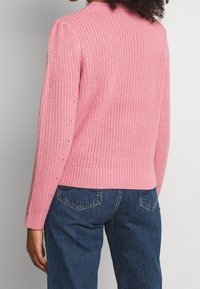 Pull rose en maille avec un motif texturé, poignets et ceinture côtelés. Porté avec un jean denim bleu, affichant une coupe décontractée.