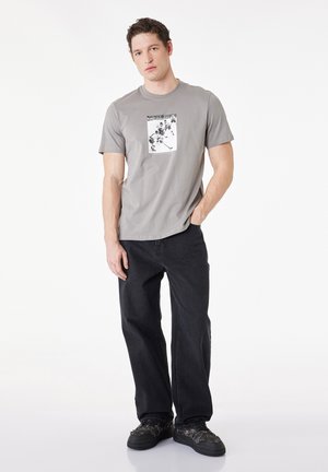 Jonge man staat met één hand in zijn zak, draagt een grijs t-shirt met grafische print, zwarte spijkerbroek en zwarte sneakers op een witte achtergrond.