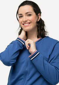 Giacca blu con zip e polsini a strisce in blu navy, bianco e rosa; texture liscia e design casual, indossata da una persona sorridente con le mani vicino al collo.
