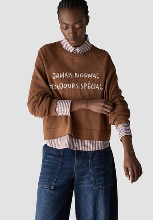 Person mit geflochtenem Haar, die einen braunen Pullover mit französischem Text "Jamais Normal Toujours Spécial" über einem gestreiften Hemd und blauen Jeans trägt.