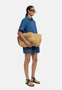 Torba plażowa z słomy o plecionym wzorze w naturalnym kolorze, z ciemnym uchwytem i ozdobnym guzikiem. W towarzystwie dżinsowej koszuli i sandałów.