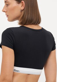 Svart kort topp laget av et mykt, elastisk materiale, med korte ermer og en hvit elastisk band med "Klein" logo nederst.