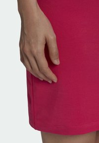 Detailní záběr na ruku odpočívající na okraji jasně růžového, hladkého texturovaného oděvu, který má lem s jemnými švy.