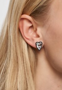 Orecchini a forma di cuore in argento con una pietra preziosa trasparente al centro. Texture liscia e finitura lucida, attaccati al lobo dell'orecchio di un modello.