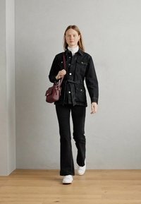 Giacca di jeans nera con cintura in vita, abbinata a pantaloni neri a campana e scarpe da tennis bianche. Una piccola borsa bordeaux è indossata a tracolla.