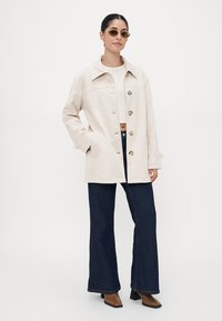 Beige oversized jacket with button closure, two side pockets, paired with dark wide-leg jeans and heeled shoes. Model wears sunglasses.