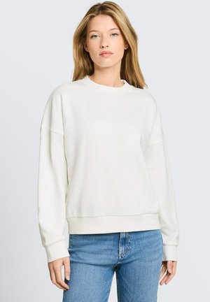 Junge Frau mit langen blonden Haaren, die einen weißen übergroßen Pullover und blaue Jeans vor einem schlichten hellen Hintergrund trägt.