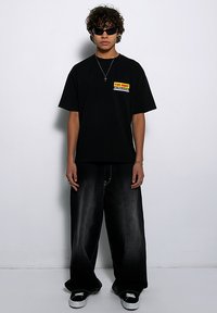 Czarny bawełniany t-shirt z wyrazistym napisem "NA WYNAJEM". W połączeniu z szerokimi czarnymi dżinsami i czarnymi trampkami. Modelka w ciemnych okularach przeciwsłonecznych.