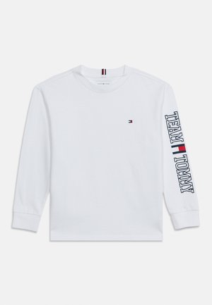 Hvid langærmet skjorte med lille Tommy Hilfiger flaglogo på brystet og "TEAM TOMMY" med logo trykt vertikalt på højre ærme.