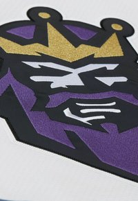 Logo ricamato in nero e viola con una corona e un volto in accenti bianchi e dorati su uno sfondo bianco testurizzato.