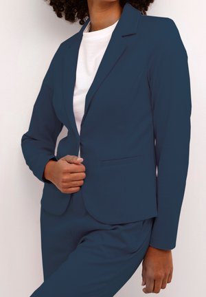 Vrouw draagt een marineblauw blazer en bijpassende broek over een wit overhemd, en past de blazer aan met één hand.