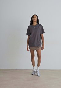 Sivi majica s diskretnim Nike logotipom, uparena s bež suknjom. Model nosi bijele čarape i srebrne tenisice, stojeći na neutralnoj pozadini.