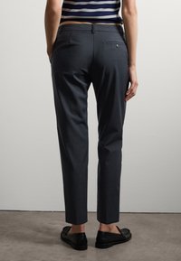 Dunkelgraue maßgeschneiderte Hose mit glatter Textur, schmaler Passform und einer Gesäßtasche mit einem Knopf. Getragen mit schwarzen Schuhen.