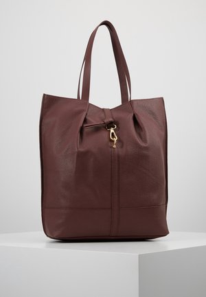 Sac fourre-tout en cuir marron foncé avec double bandoulière et fermeture à fermoir doré au centre supérieur, présenté sur un piédestal blanc.