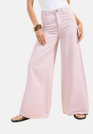 Kobieta ubrana w jasnoróżowe spodnie z szerokimi nogawkami, biały top, czarne sandały oraz brązowe bransoletki z koralików na lewej ręce, stojąca ze swobodnie opuszczonymi rękami.