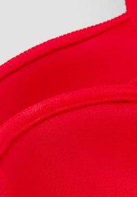 Soutien-gorge rouge en tissu lisse, présentant une forme sculptée. Il a un design sans coutures et une bordure robuste et texturée le long des bords.