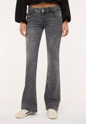 Donna che indossa jeans grigi a zampa con vita a doppio bottone e sneakers bianche, in piedi davanti a uno sfondo neutro.